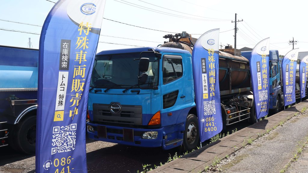 屋外に駐車された複数の青い特殊車両(タンクローリーなど)と、手前に等間隔で設置された青いのぼり旗の写真。のぼり旗には上部にTMCのロゴがあり、「特殊車両販売中」の大きな文字、「在庫検索 トムコ 特殊」という検索ボックス、スマートフォン読み取り用のQRコード、電話番号(086-441-8400)がプリントされています。